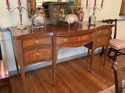 A serpentine mahogany sideboard set with silver candelabra, a shell-shaped dish, and a tole tray in a dining room